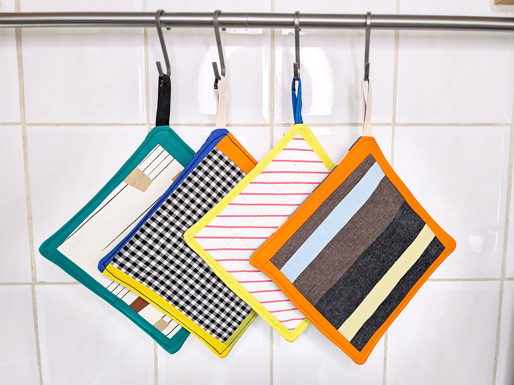 Quatre maniques de couleurs différentes (vert, jaune, orange, vichy et multicolore) accrochées sur une crémaillère.